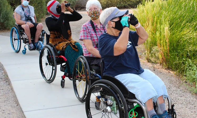 Four adults using wheelchairs travel along a paved outdoor path, with two looking through binoculars.