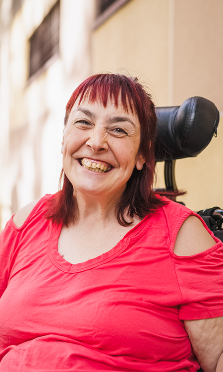 A smiling woman with red hair sits in a wheelchair outdoors wearing a bright red shirt.