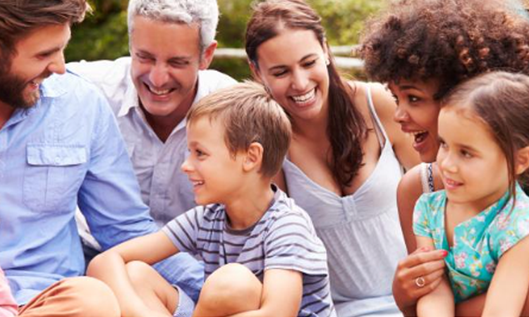 A diverse group of adults and children sit close together outdoors smiling and laughing in a joyful moment
