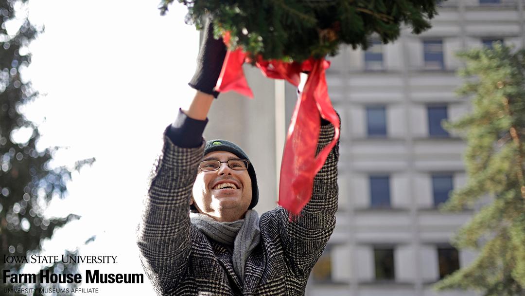 Nicholas Scott smiling while hanging a wreath outside at the Farm House Museum. Graphic says Iowa State University Farm House Museum University Museums Affiliate