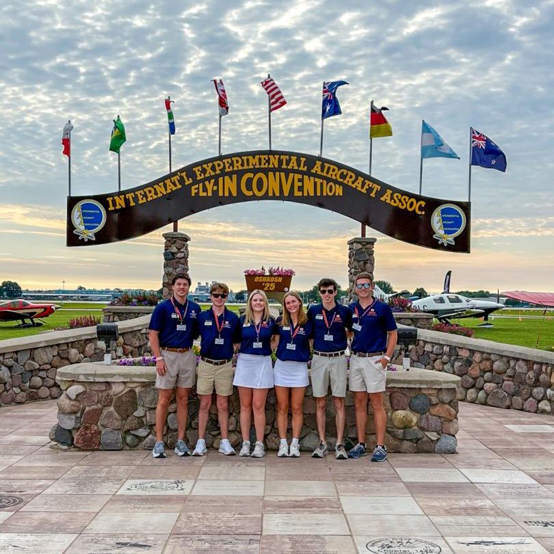 Six Stripe Wing Aviation Ambassadors stand in front of the Osh Kosh sign