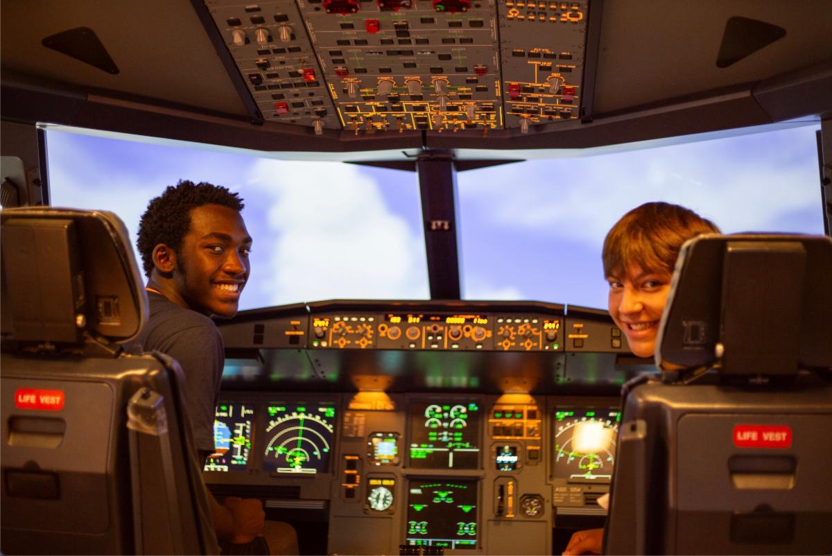Two boys sit in the cockpit of an AirBus simulator while smiling.