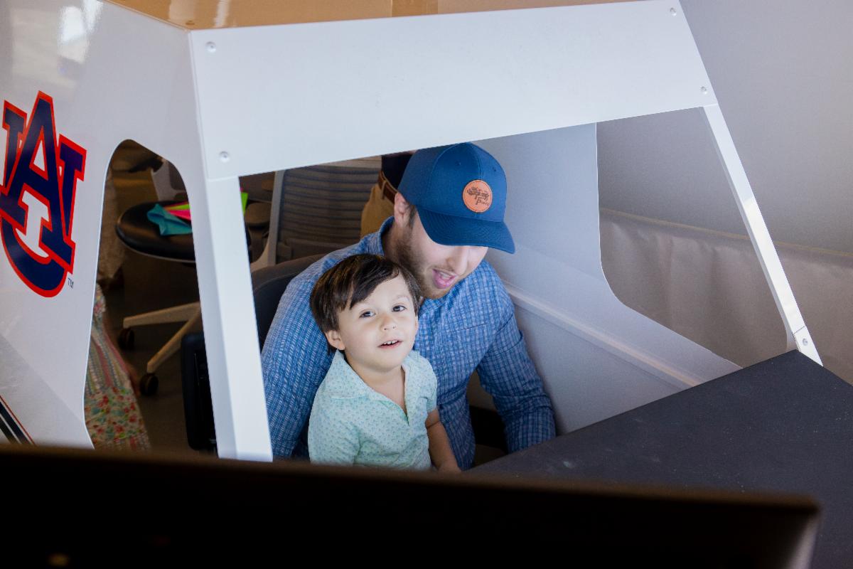 Young child in father's lap in looking up at camera while in airplane simulator.