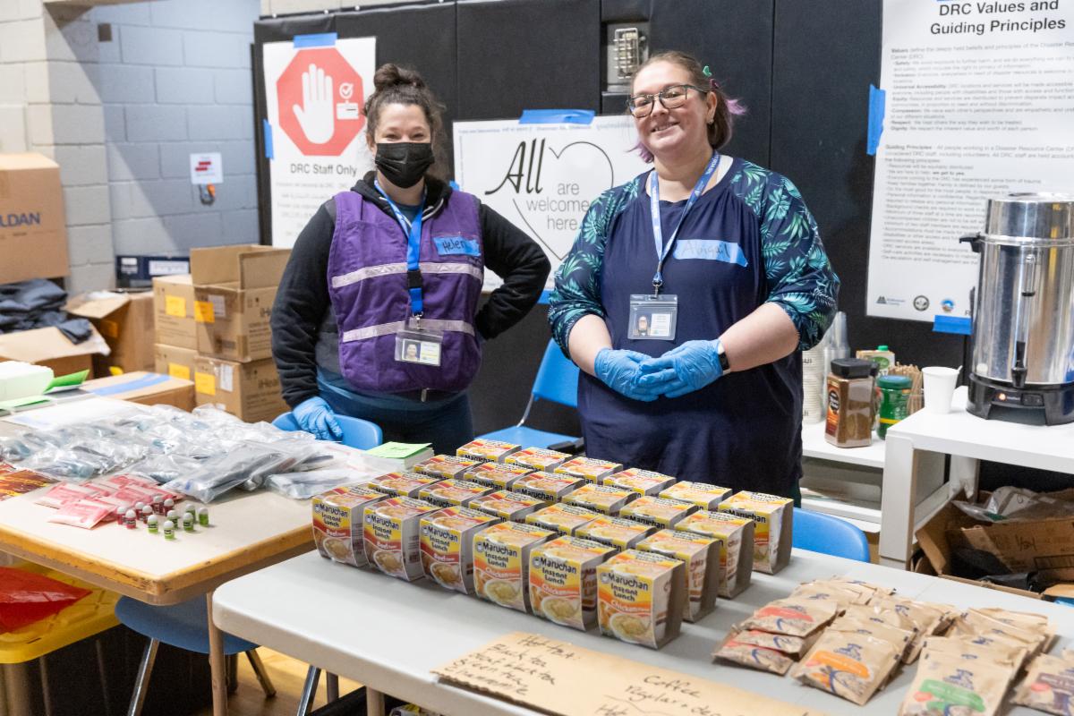 Two shelter staff stand behind a table of snacks and hygiene items, smiling invitingly. Behind them is a sign that reads "All are welcome here." Imago Dei, January 2024. 