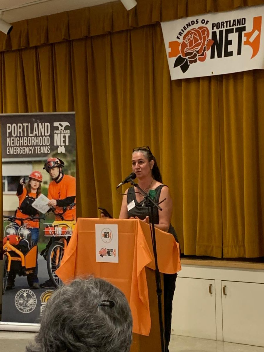 Alice Busch stands behind a podium at a Portland NET event, speaking to an audience.
