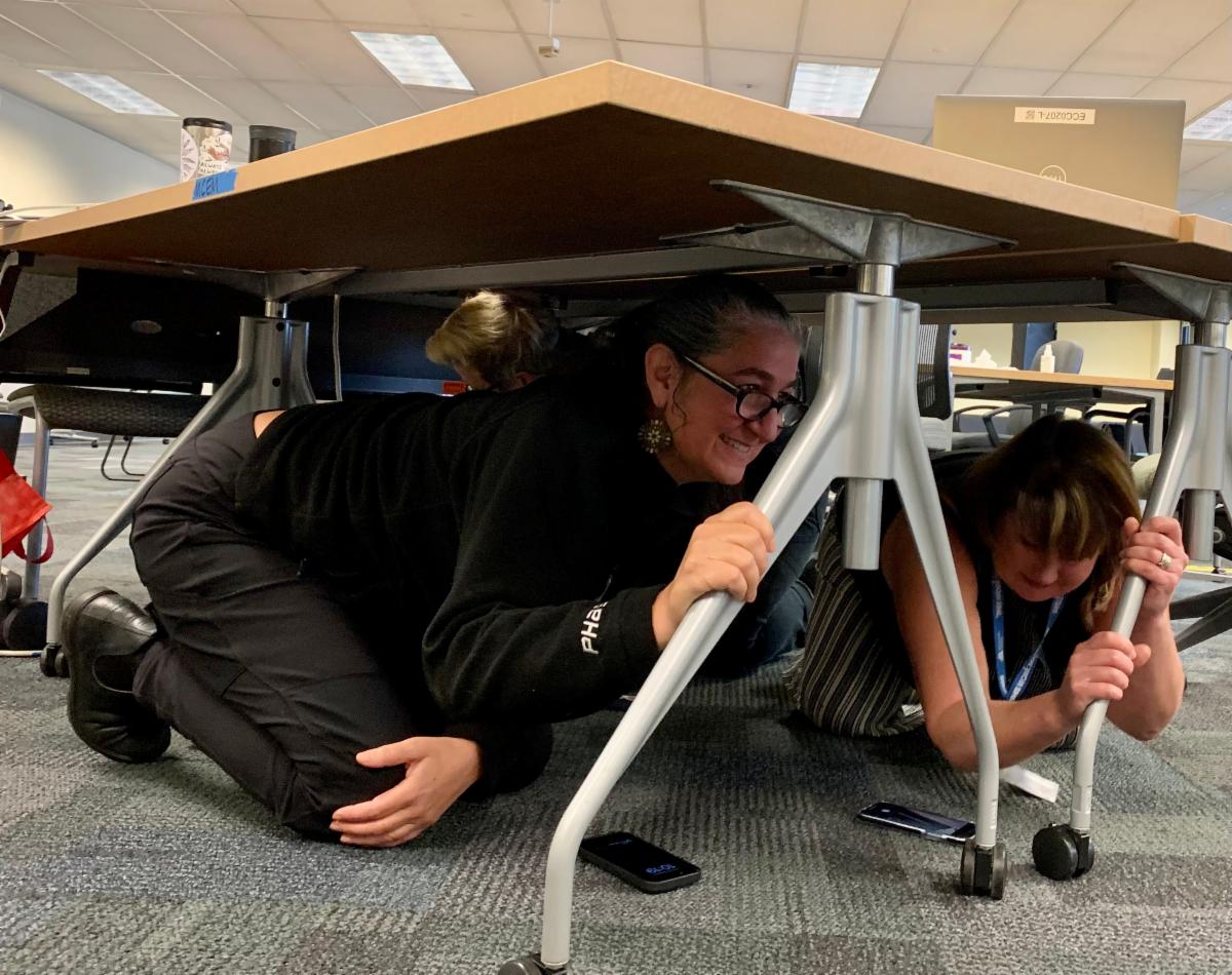 Kirsten Kuppenbender, Alice Busch and Allie Callow-Spencer on the ground, holding on to table legs underneath the table.
