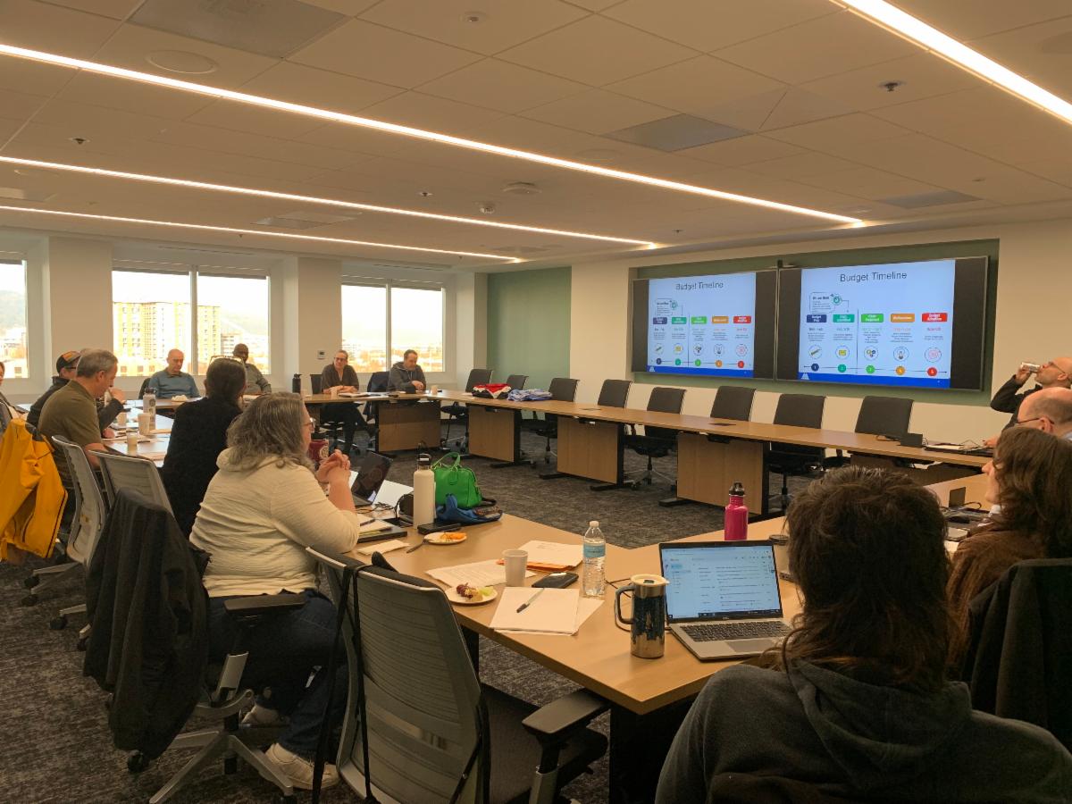 MCEM Staff sit at a U-shaped table with presentations displayed on two screens.