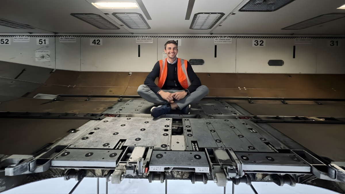 A man in an orange safety vest sits, smiling, in a large empty aircraft baggage area.