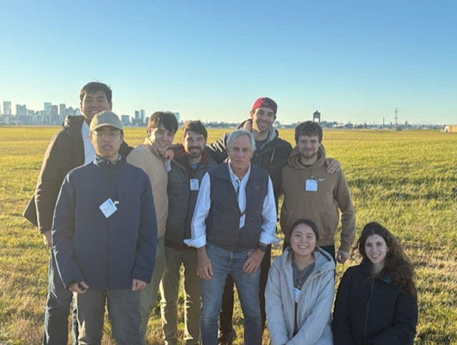 A group of nine people posing in a grassy field with the Boston skyline in the background.