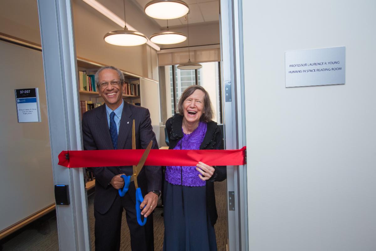 A photo of Daniel Hastings and Vicki Young standing in the doorway of the Laurence Young Humans in Space Reading Room. A large red ribbon stretches across the door frame in front of them. Dan holds scissors, about to cut through the ribbon.