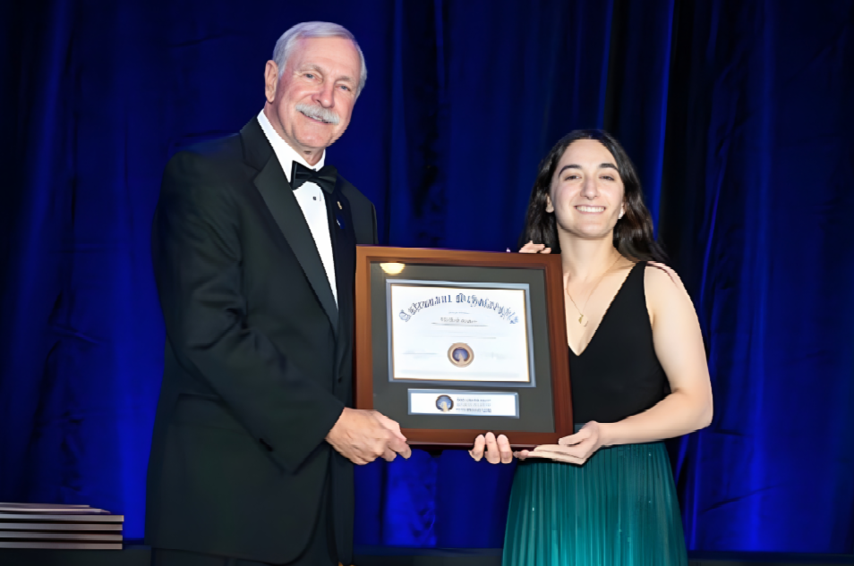 Two people holding a framed award in front of a blue curtain backdrop.