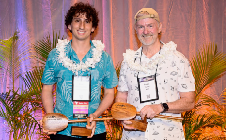 Two men wearing leis hold wooden paddle-shaped awards in front of a palm leaf and curtain background.