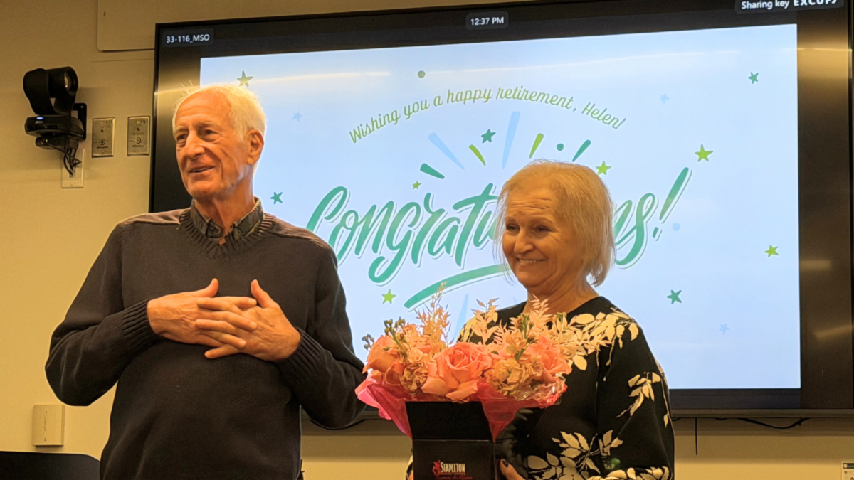 Two people at a retirement celebration, with a congratulatory message on a screen and a bouquet of flowers.  Transcribed Text:  "Wishing you a happy retirement, Helen! Congratulations!"