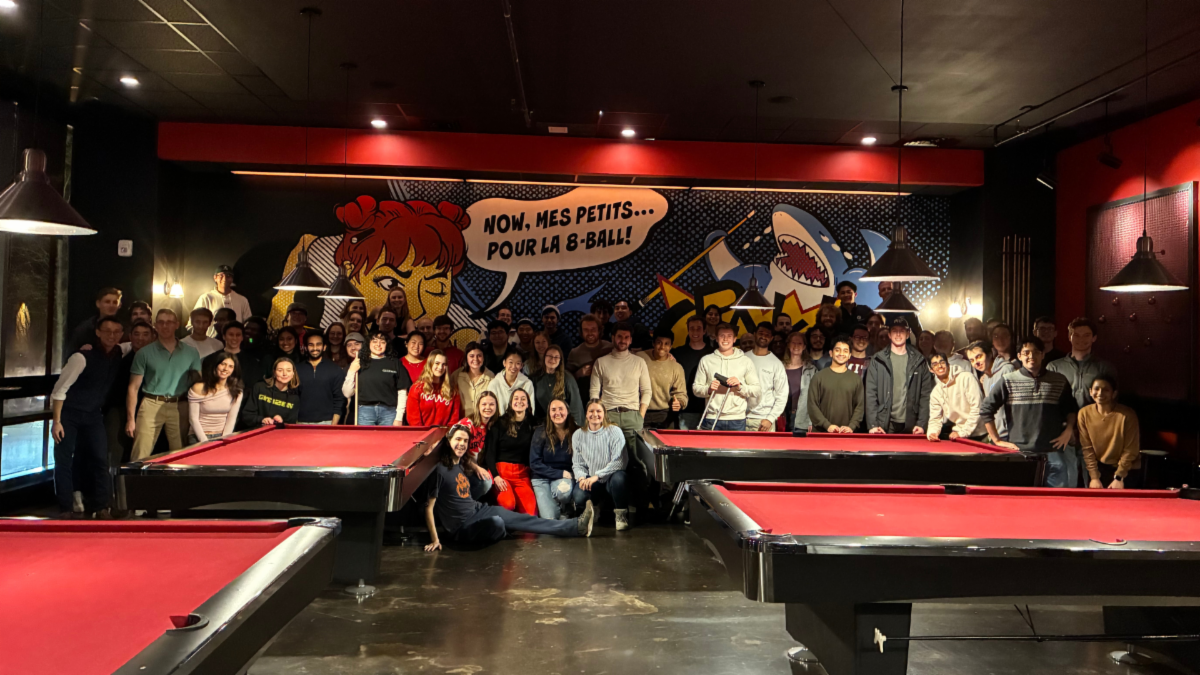 A large group of people posing in front of pool tables, with a pop art mural of a person and a shark in the background.  Transcribed Text:  "NOW, MES PETITS... POUR LA 8-BALL!"