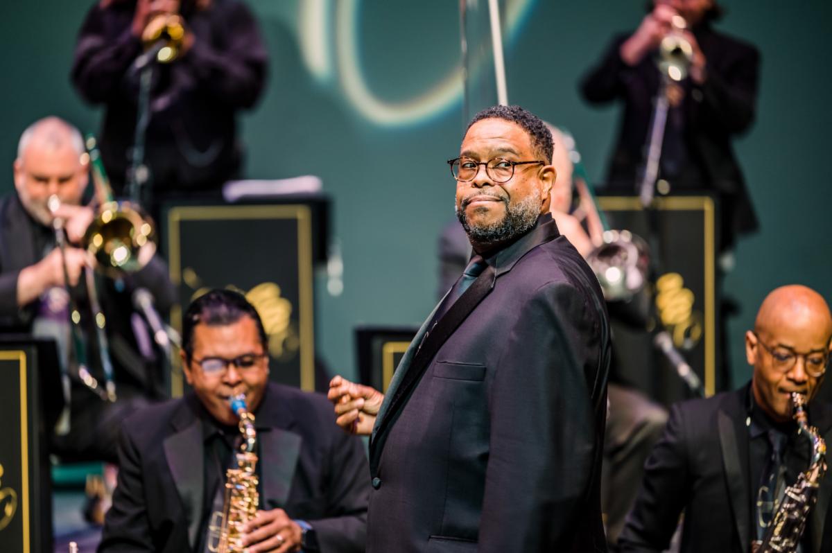Jazz orchestra performing on stage in black suits with black shirts and ties the conductor is in front of the two saxophonists one trombonist and two trumpet players