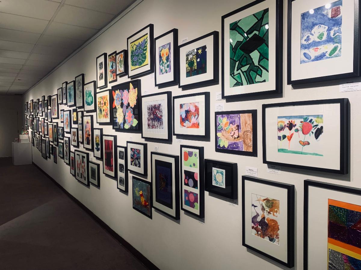 Numerous pieces of artwork created through the Art Works for All program hung on a white wall for an exhibition