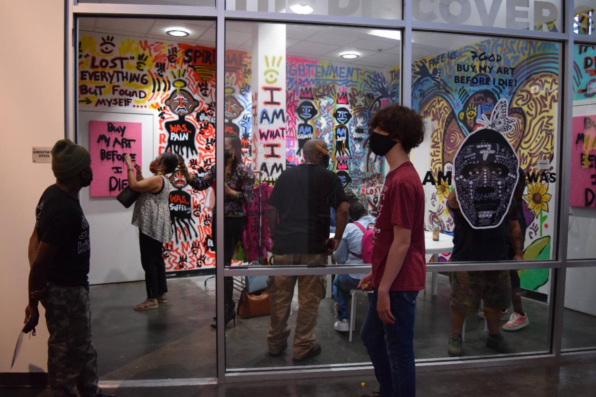 People looking at an art exhibit at the Delaware Contemporary of a room filled with colorful graffiti-like paintings and a clothing rack that is part of the exhibit