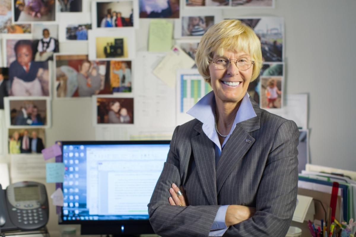 Portrait of Terry Miller standing with her arms crossed over her chest in an office with a desk holding a computer monitor and files and a wall covered in photos in the background