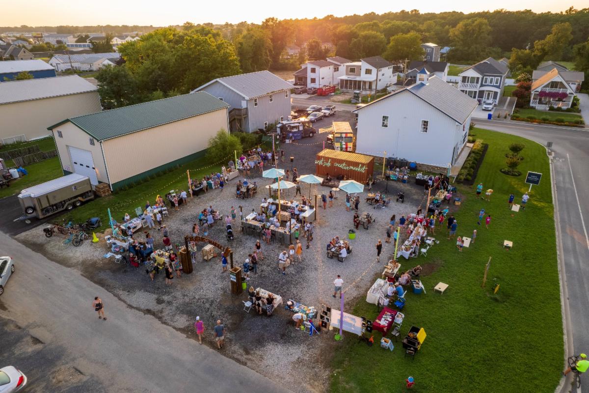 Overhead shot of Creative Market West Rehoboth showing vendor tables and food trucks laid out on an asphalt lot with people all around the market the lot is surrounded by buildings and a strip of grass that has lawn games on it