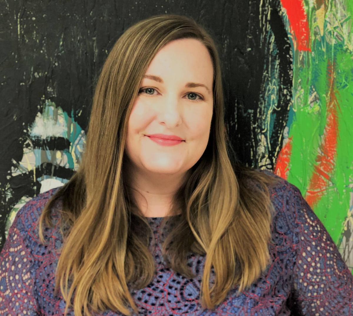 headshot of Jessica Ball Director of the Delaware Division of the Arts looking straight at the camera while standing in front of a piece of art that fills the whole background of the picture