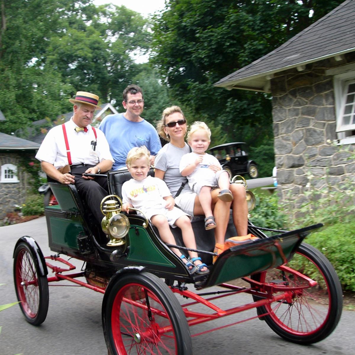 Family unit of 4 riding in a green and red antique car with a man in period clothes driving the car