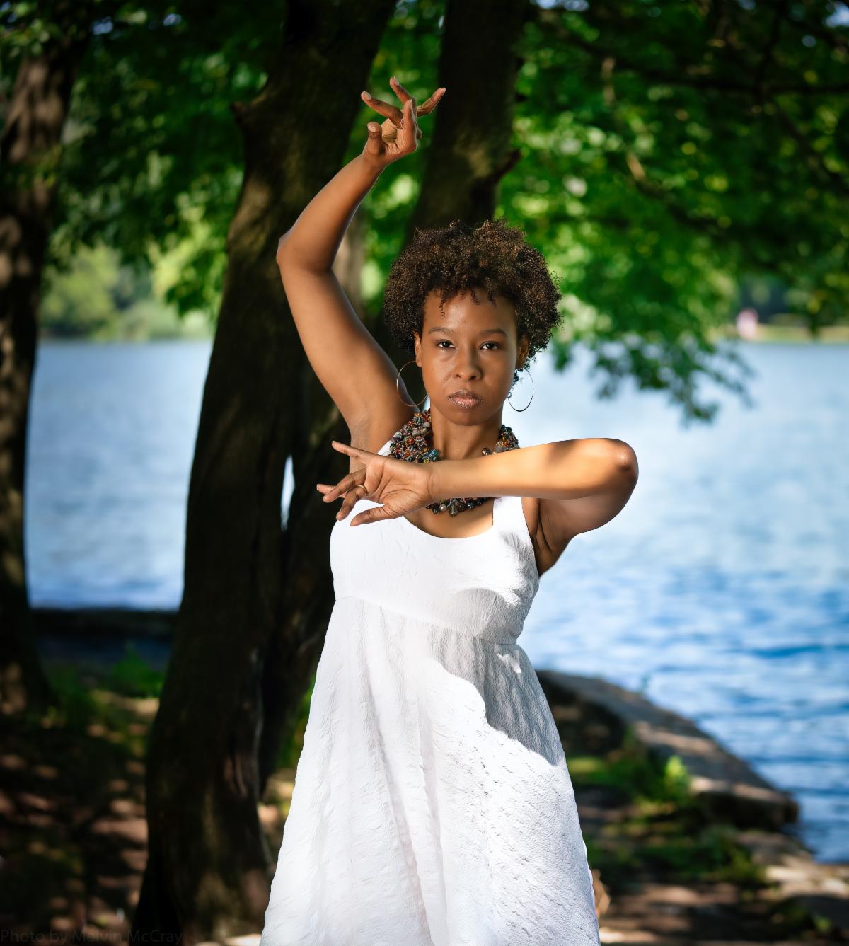 three quarter portrait of Dr Jame McCray in mid dance with one arm stretched above her head and the other crossed in front of her chest outside in front of a tree and a body of water