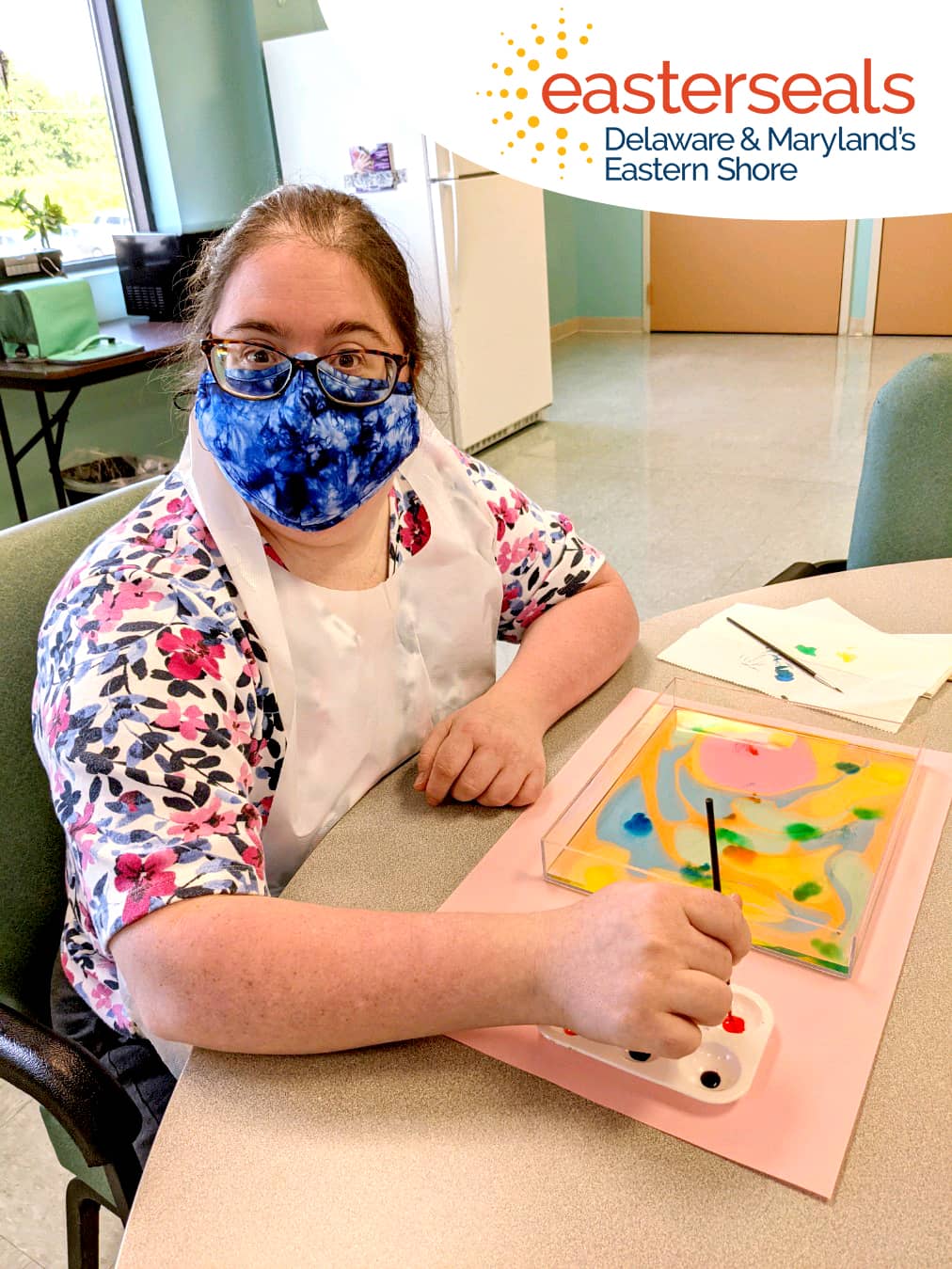 An Artfulness participant is sitting in a rec room in an Easterseals facility at a table paused from working on her painting with paintbrush in hand in order to look at the camera for the photo