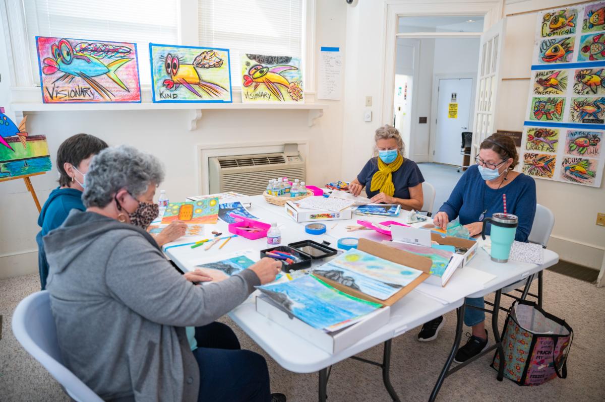 4 woman sitting around 2 folding tables pushed together to create a larger square table 2 to a side talking and creating colorful artwork with pastels in a community classroom in a Delaware public library as part of a Creative Aging program