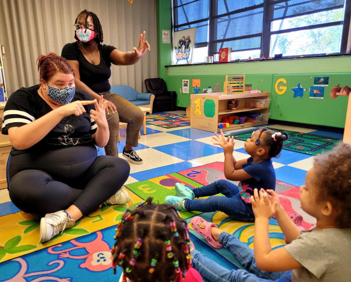 Two female adults sit in front of three young female children showing the children how to do a motion with their hands while the children imitate