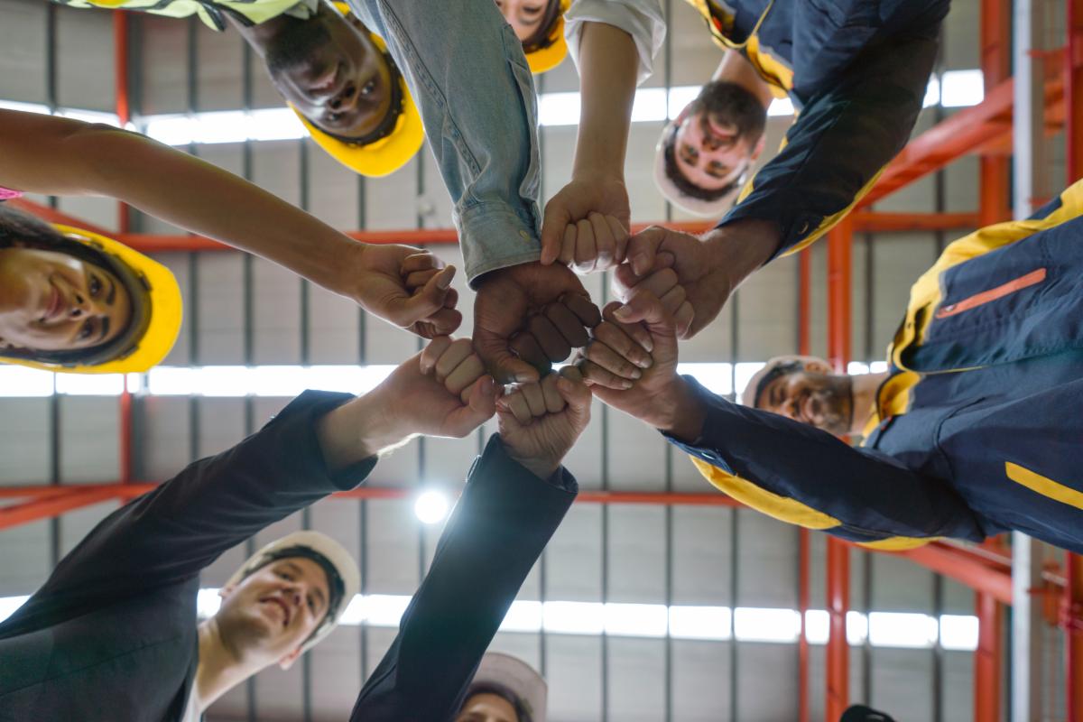 Group of male and female factory labor bump their fist together after finish meeting. Everyone wearing safety uniform and helmet. Workers working in the metal sheet factory. Bottom View