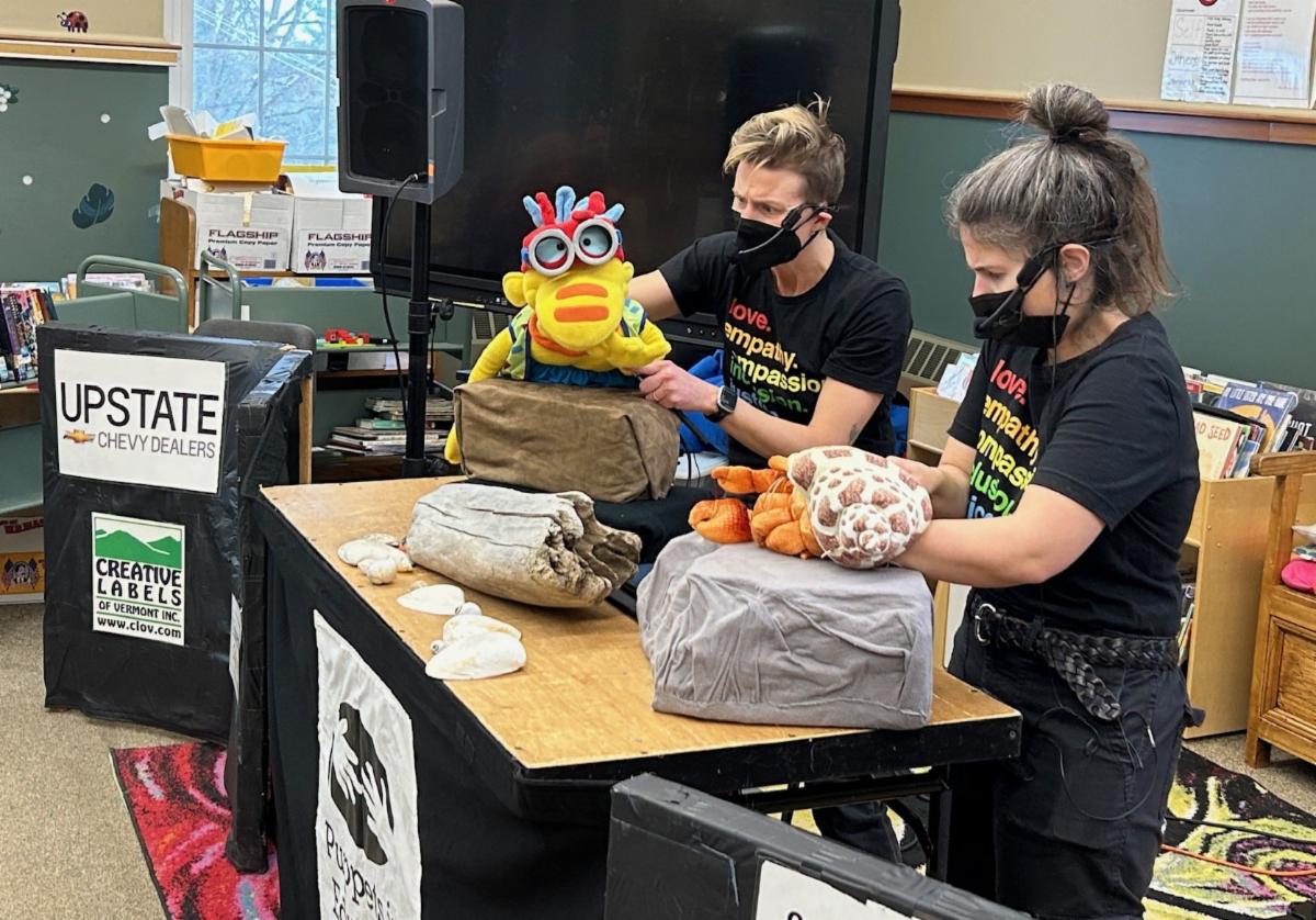 Two puppeteers in wearing black in front of a classroom perform the Resilience Puppet Program featuring a hermit crab puppet