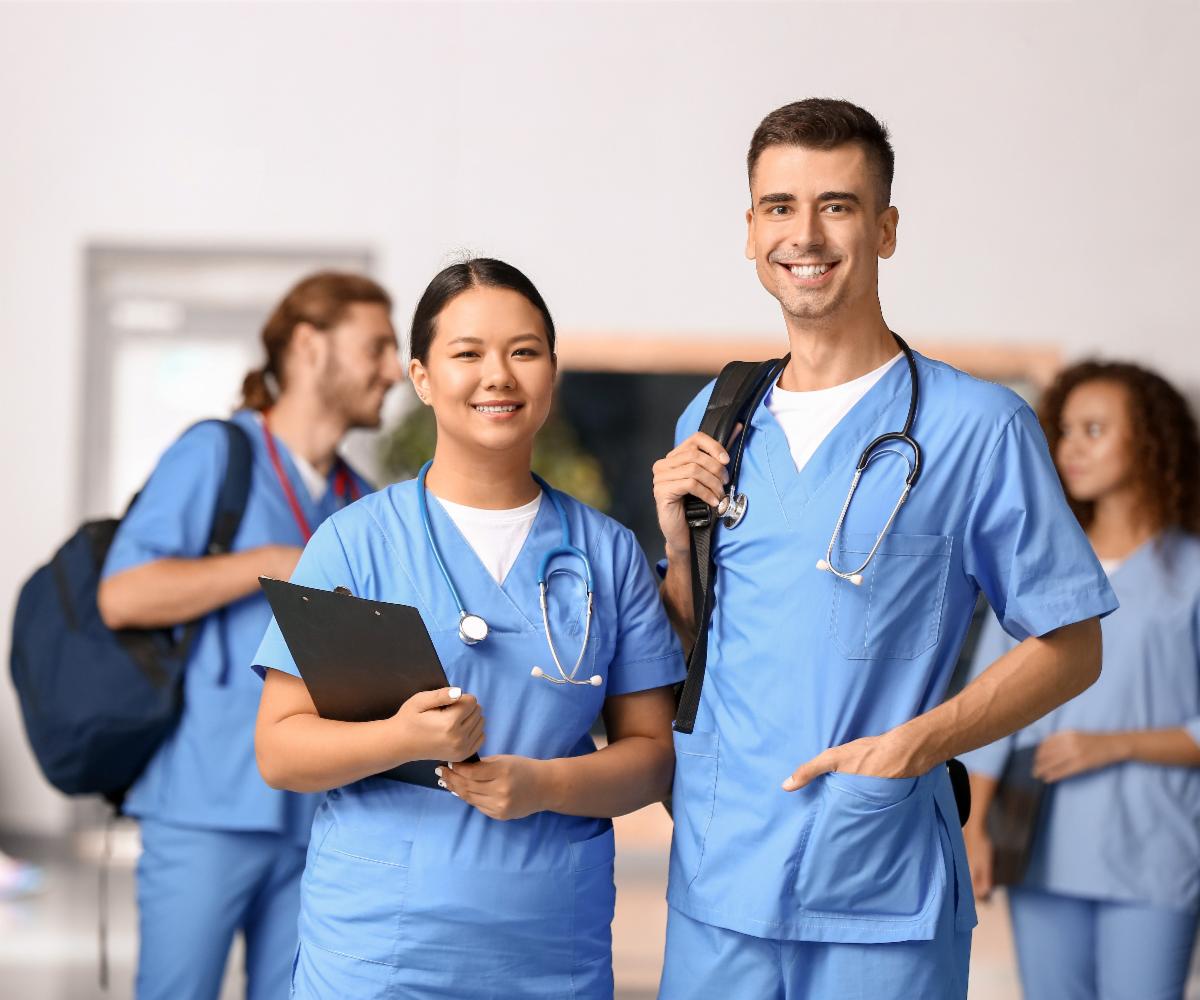 A group of smiling medical students wearing blue scrubs and carrying notebooks and backpacks stand in a college hallway
