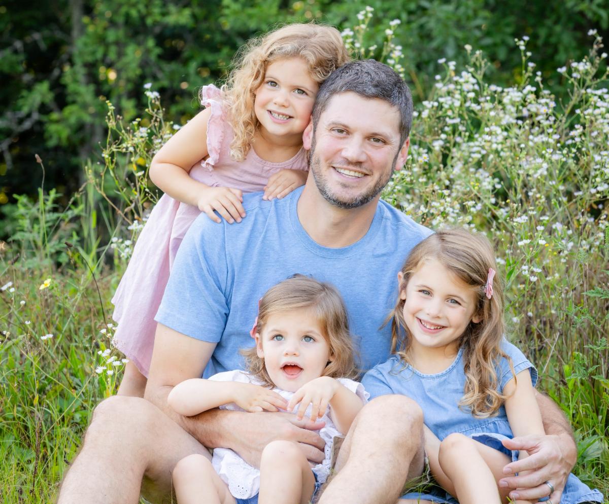 A father weraing a blur t-shirt sits on the grass with his three children wearing blue, pink and white and all are hugging