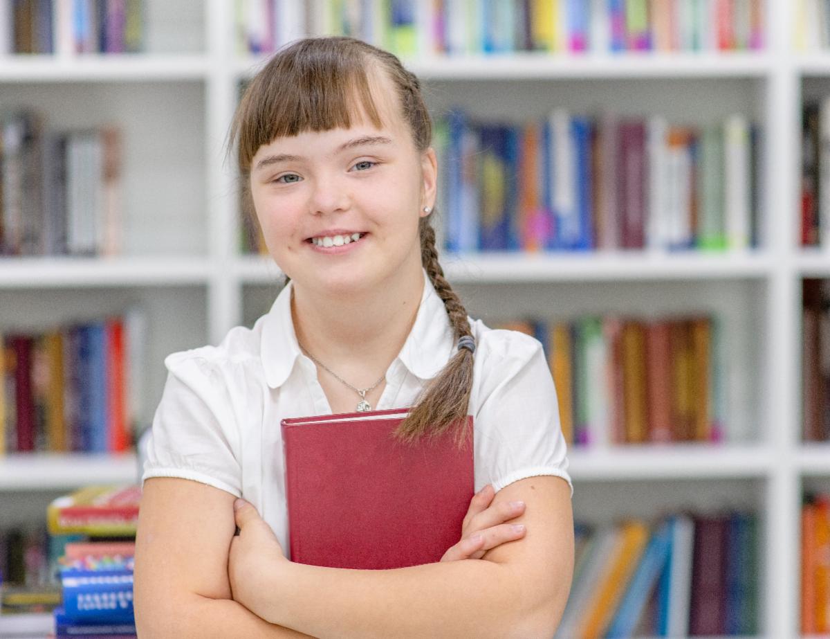 A student with hair in braids and wearing a white shirt holds a red book and stands in front of a shelf of books