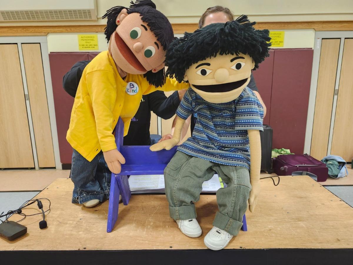 A photo of the puppet Melody, wearing yellow shirt, putting an arm around the puppet Simu, wearing a striped shirt, who is sitting on a chair.