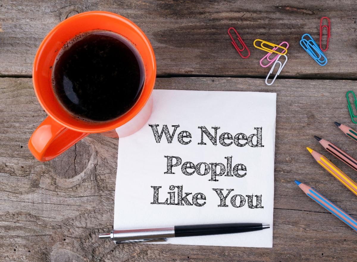 An photo looking down at a table top with an orange mug of coffee, paper clips, pens and a note that reads "we need people like you"