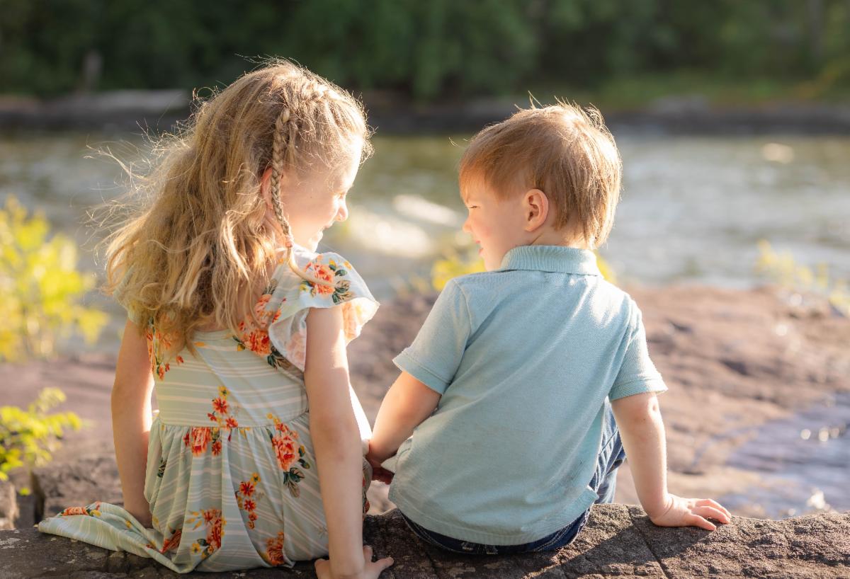Two smiling children backlit by sunlight sit on rocks by the lake with their backs toward us