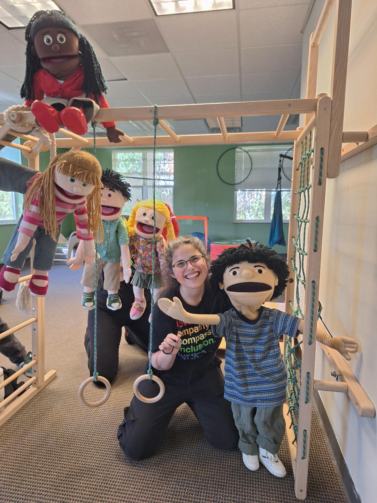 A picture of Jillian smiling while surrounded by five puppets playing on a wooden jungle gym above her