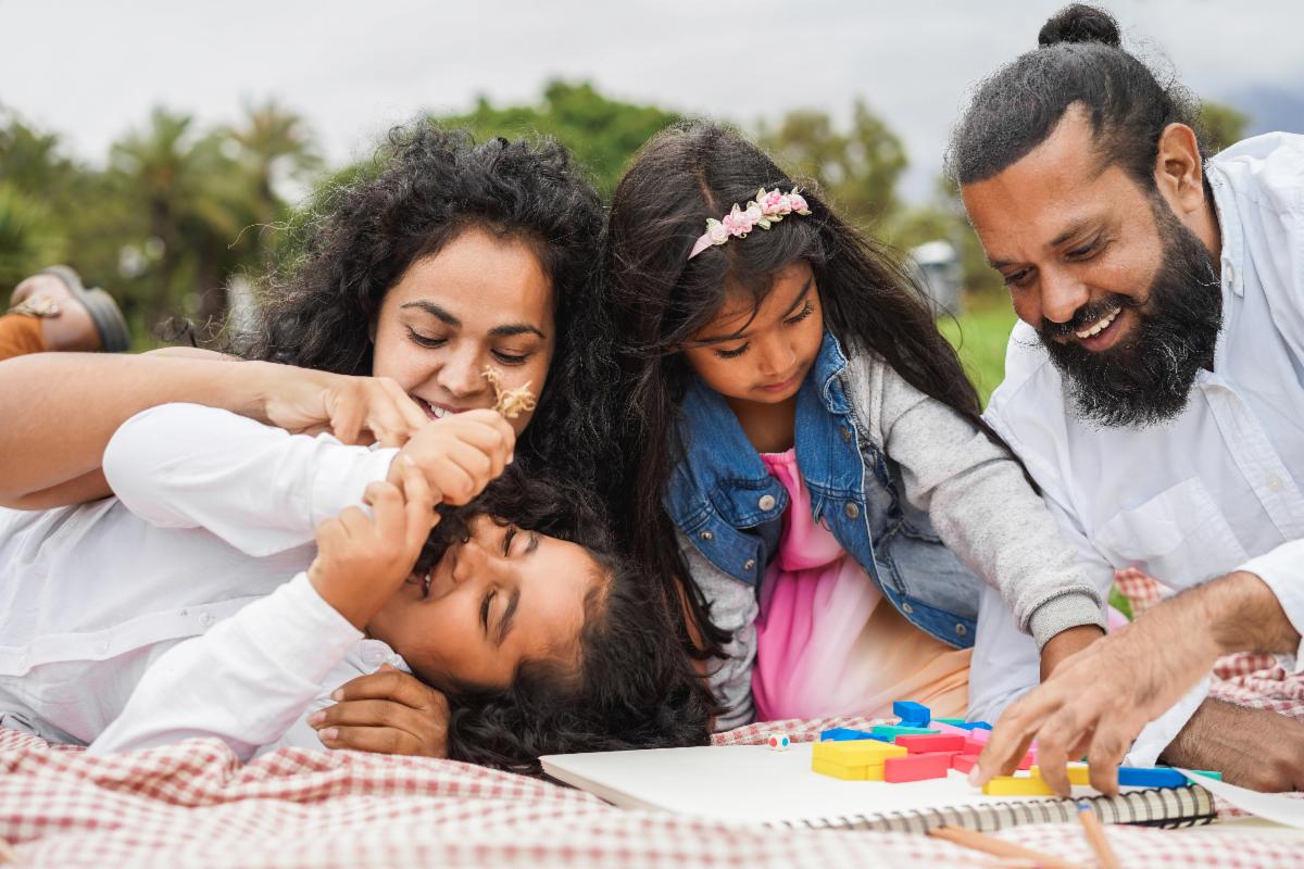 A smiling family lays on a picnic blanket while doing a puzzle and silly wrestling