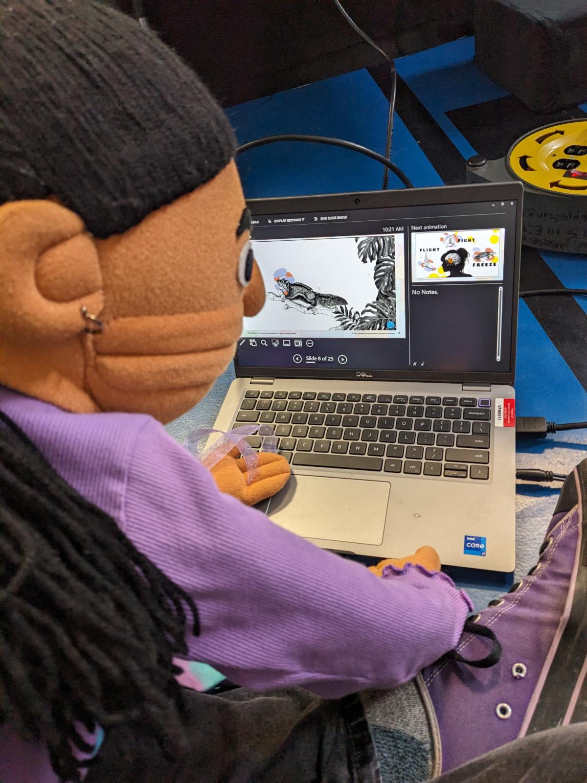A puppet wearing a purple shirt sits at a table looking at a laptop perhaps scheduling a puppet show