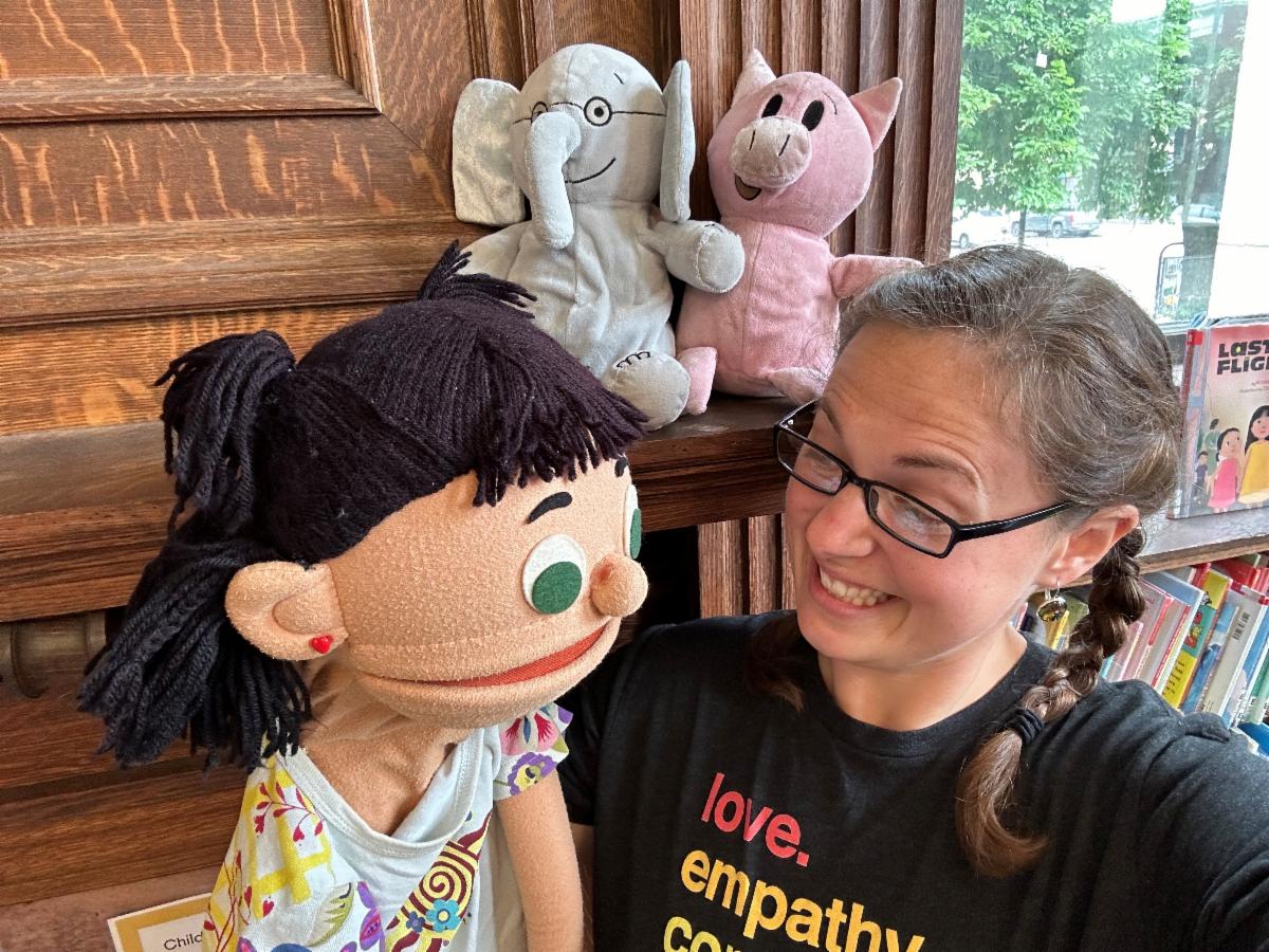 A puppeteer with dark hair sitting in front of a library bookshelf, wears glasses and a black t-shirt with the word "love," and talks to a puppet with dark hair and a flowered shirt 