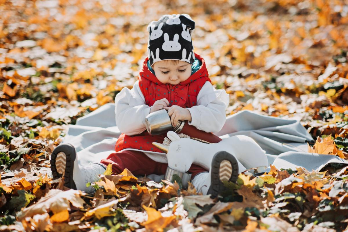A young child in a white snowsuit and red scarf sits on a blanket in the middle of autumn leaves while their stuffed unicorn drinks from a thermos cup