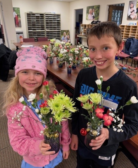 Two smiling Sibshop youth hold up flower vases filled with white, pink and lime green flowers
