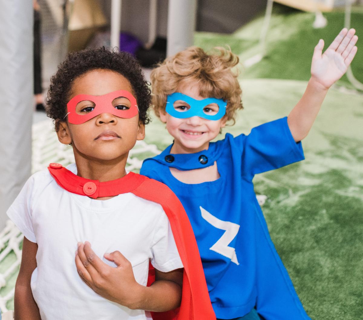 Two young children pose dressed in superhero costumes. One wears a red cape and mask and the other wears a blue shirt cape and mask.