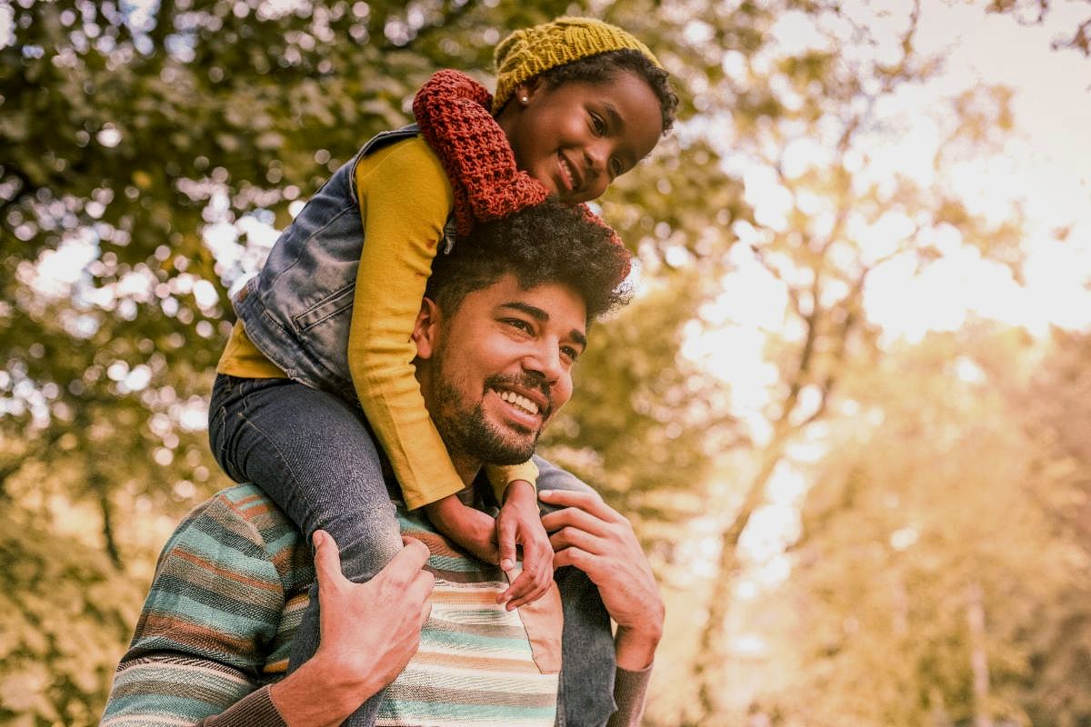 A smiling parent holds a smiling child on their shoulders while walking in the sunshine by some trees