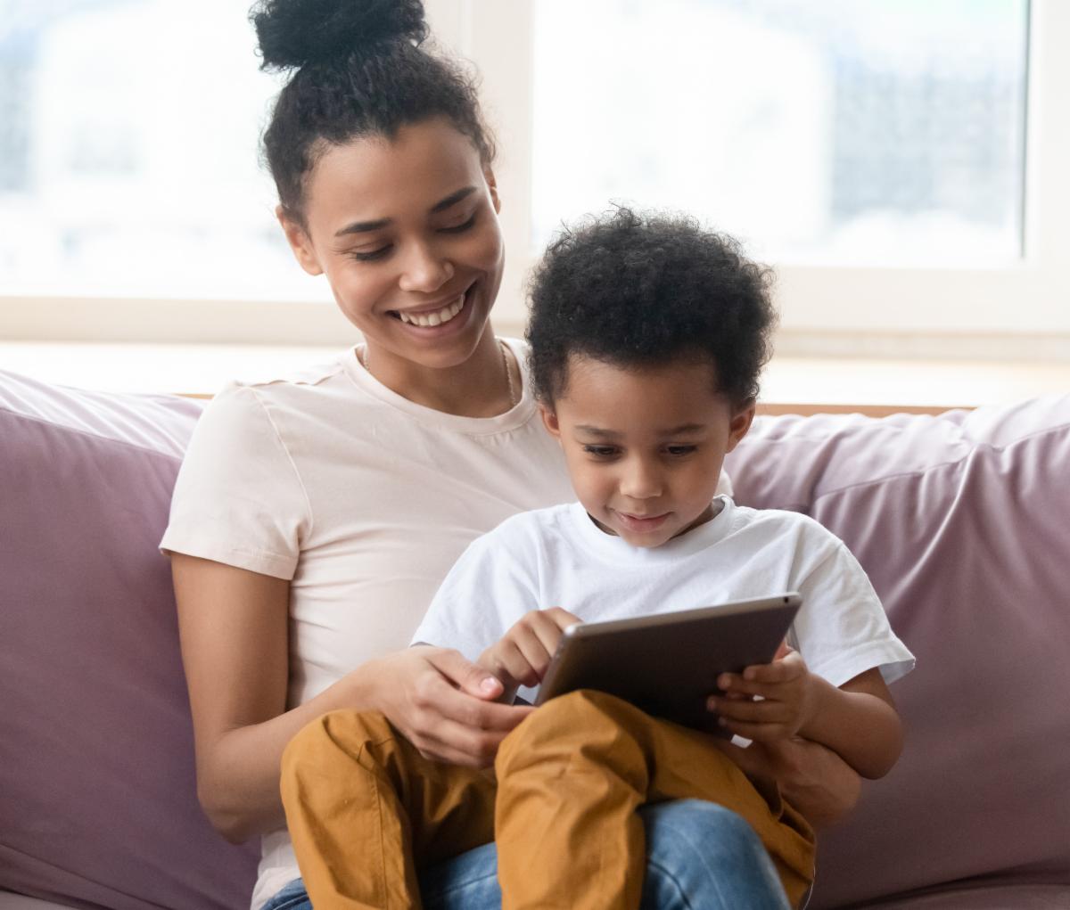 A parent sits on the couch with a child in their lap and both are looking at an iPad