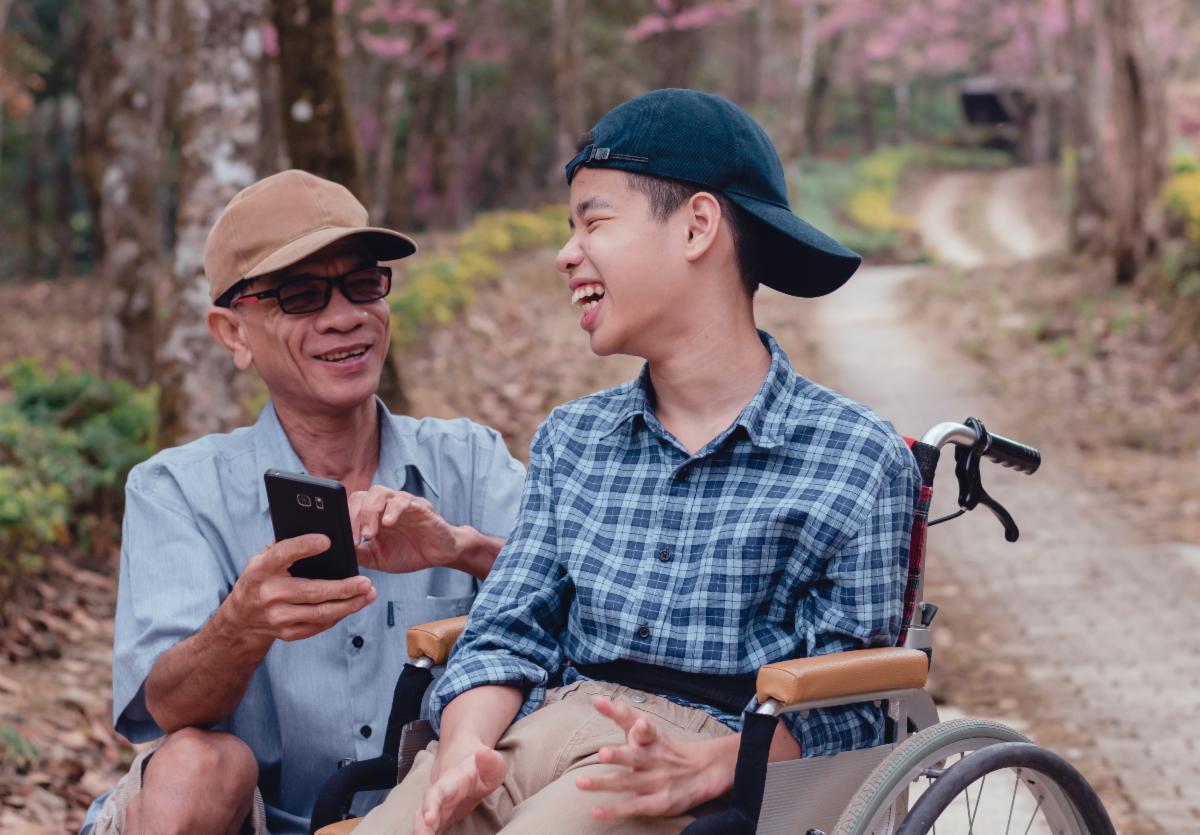 Parent wearing sunglasses and a baseball hat, laughs with a youth wearing a backwards baseball hat, while sitting in a wheelchair. Both appear to be on a hiking trail in the woods.