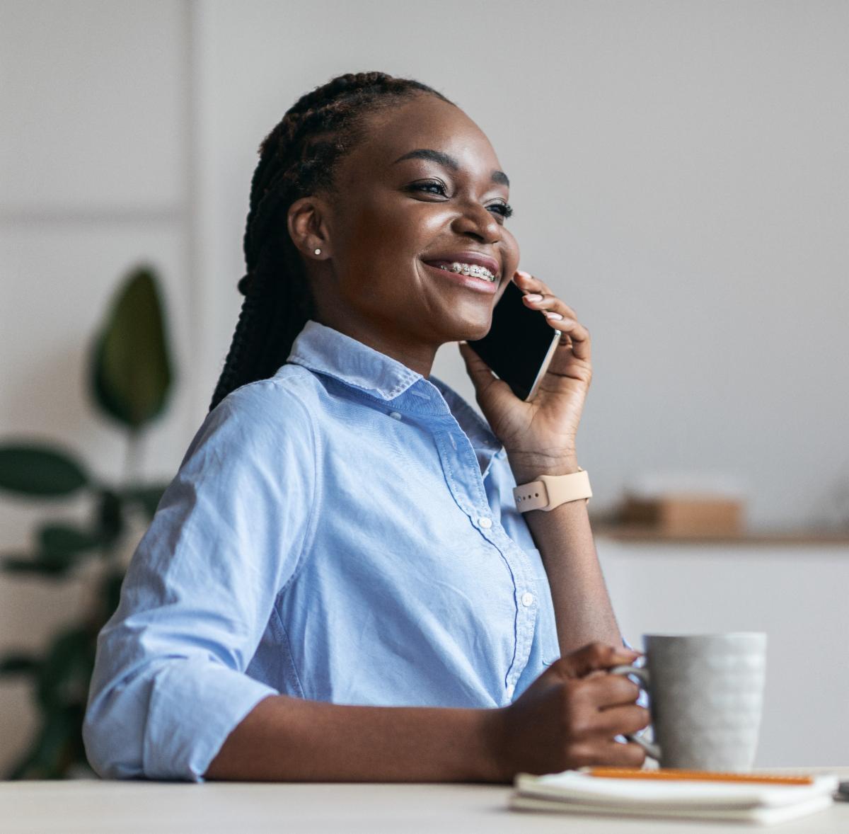 A young parent in a light blue shirt talks on the cell phone while a cup of coffee sits on the table nearby