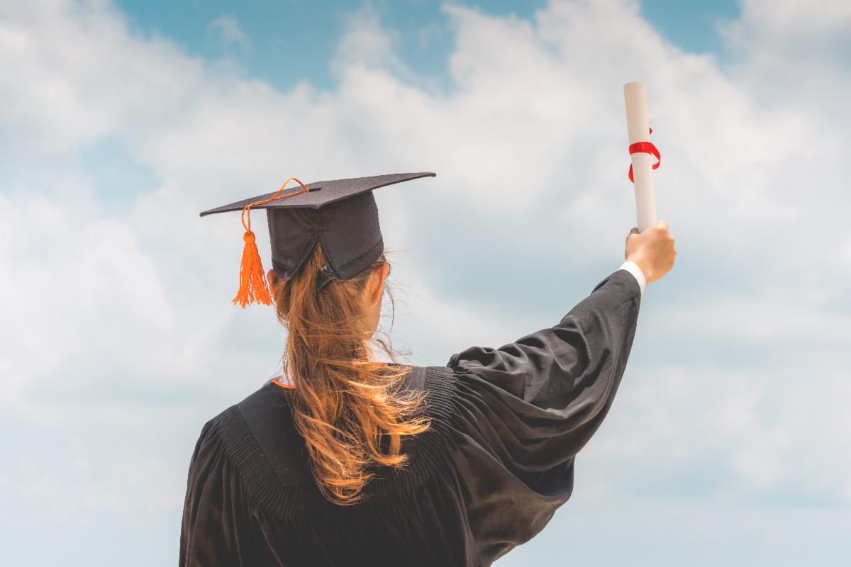 A student with back turned to us, wears a black graduation cap and gown while holding a diploma up toward a blue sky