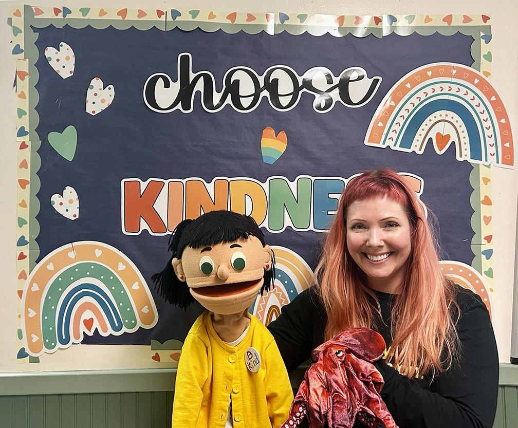 Puppeteer, Sarah and a puppet wearing a yellow shirt smile in frront of a sign that says Choose Kindness